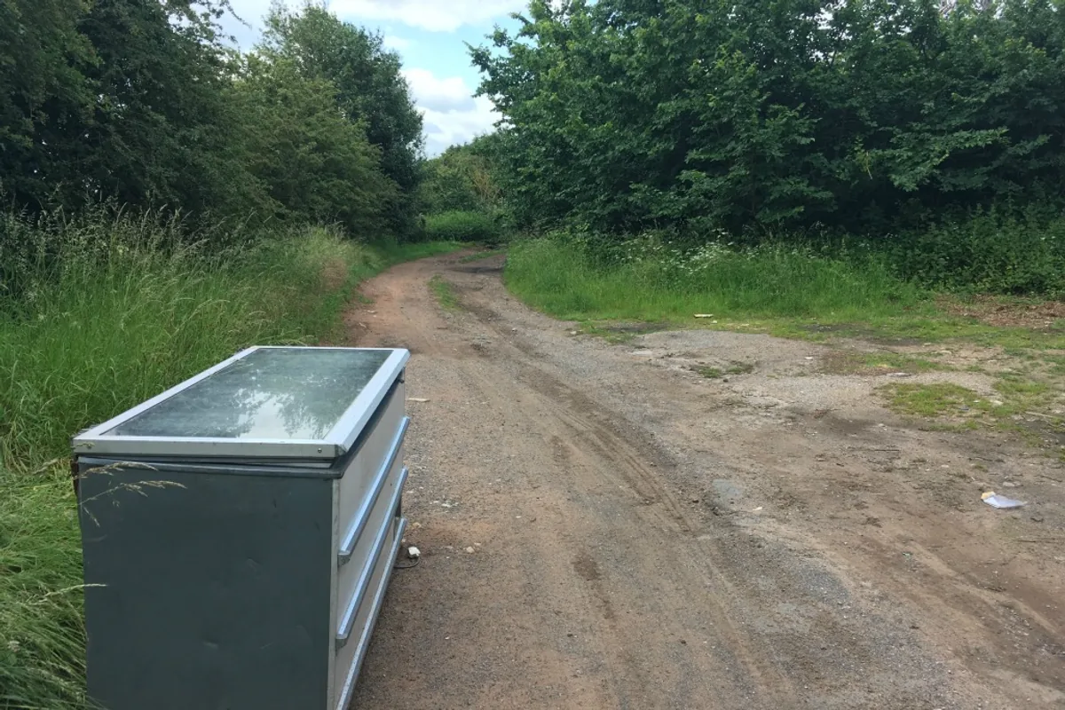 An abandoned chest freezer left standing on open ground beside a rough track, with its lid closed and its casing weathered.