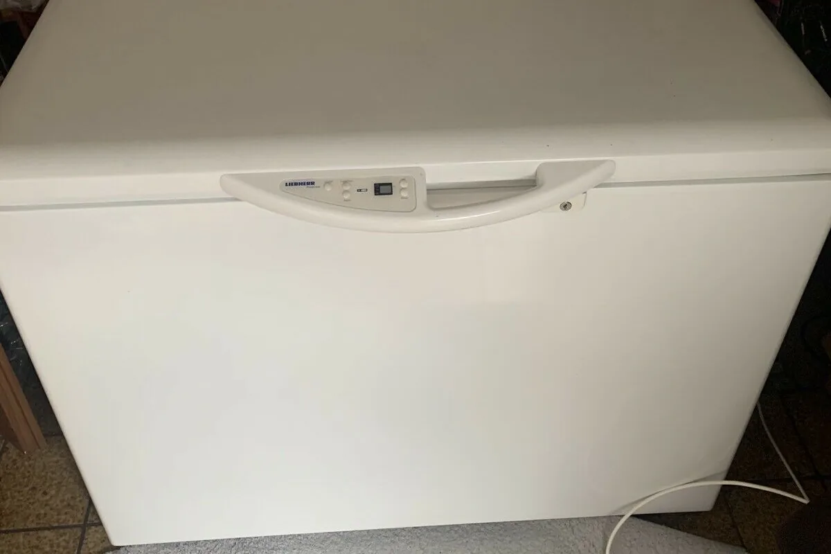 A white chest freezer standing against a wall in a domestic store area, with cardboard sheets propped behind it and its power lead trailing across the floor.