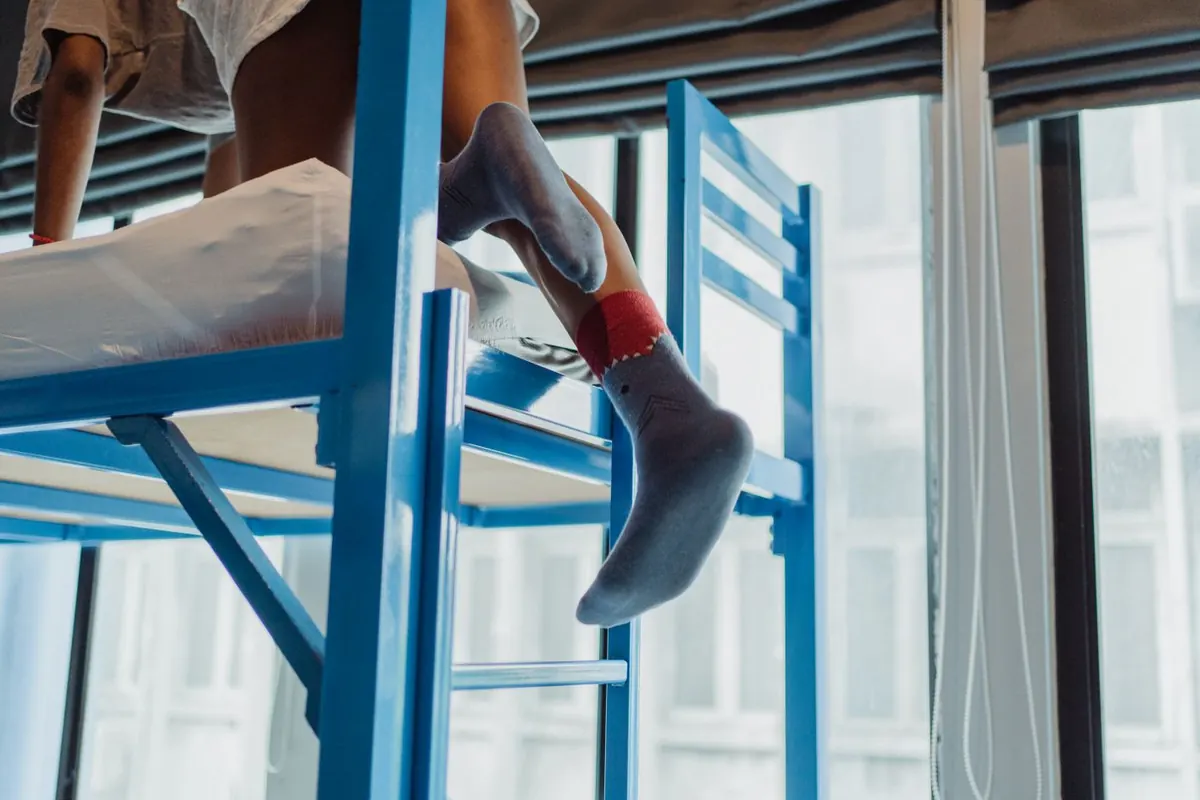 A child in socks climbing down the rungs of a blue tubular metal bunk bed ladder, with the upper mattress platform and the slatted guard rail of the frame visible above.
