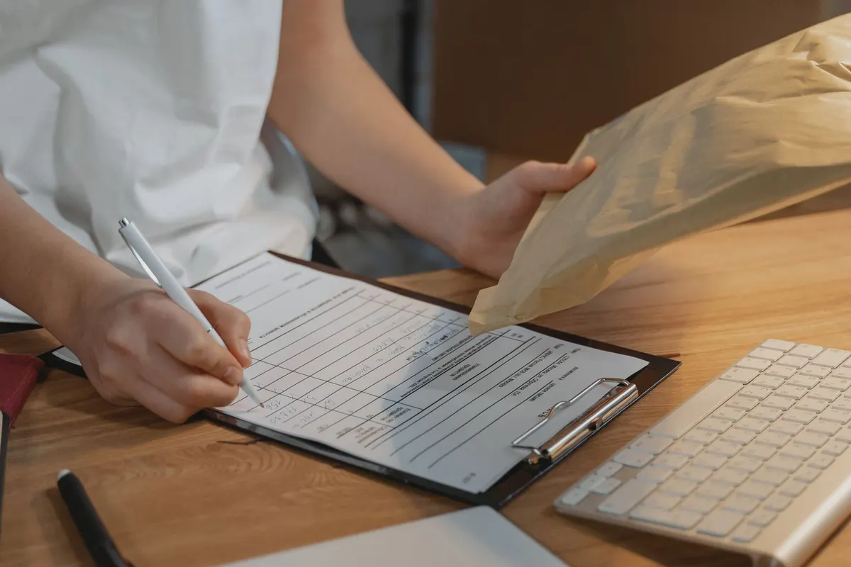 A person filling in a form on a clipboard at a desk while holding a paper envelope beside a keyboard.