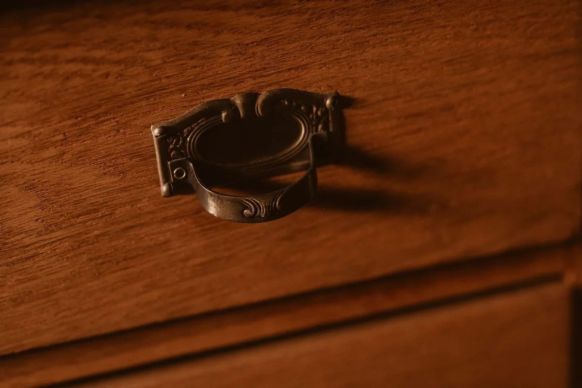 A close-up of an ornate brass drawer pull with an engraved oval backplate, mounted on a wood-grain drawer front.