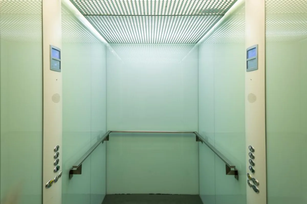The empty interior of a compact passenger lift car with frosted glass walls and a stainless steel handrail.