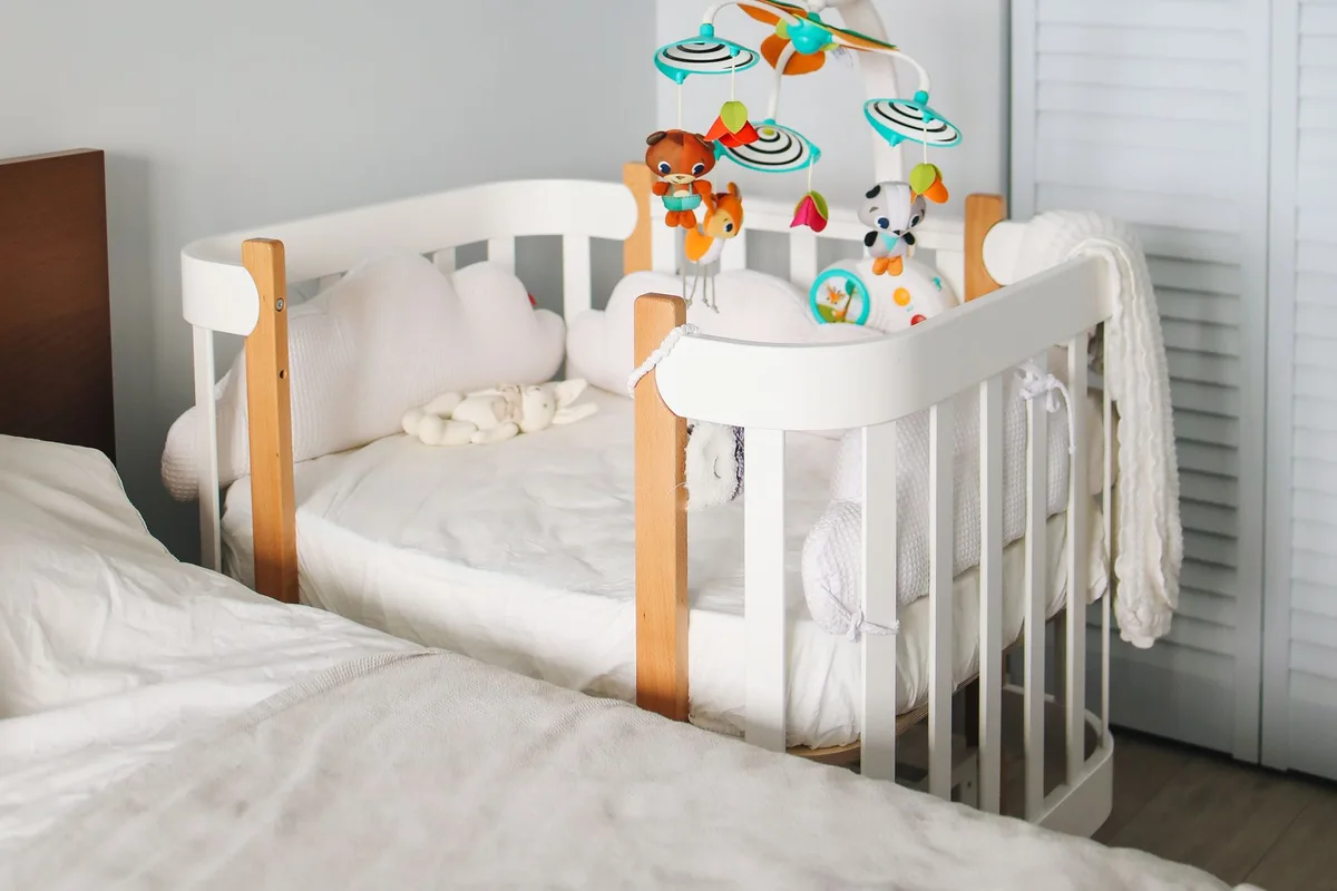 A white and timber cot with a hanging mobile toy, positioned beside an adult bed and a louvred wardrobe in a shared bedroom.