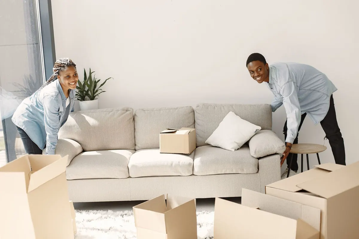 Two people lifting a beige three-seat sofa in a living room surrounded by open cardboard packing cartons.