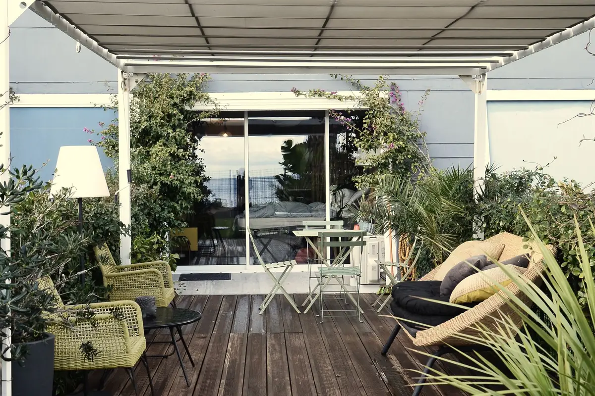 A decked terrace shaded by a fixed white awning frame, with woven armchairs, a folding bistro set and a wicker daybed arranged around a sliding glass door.