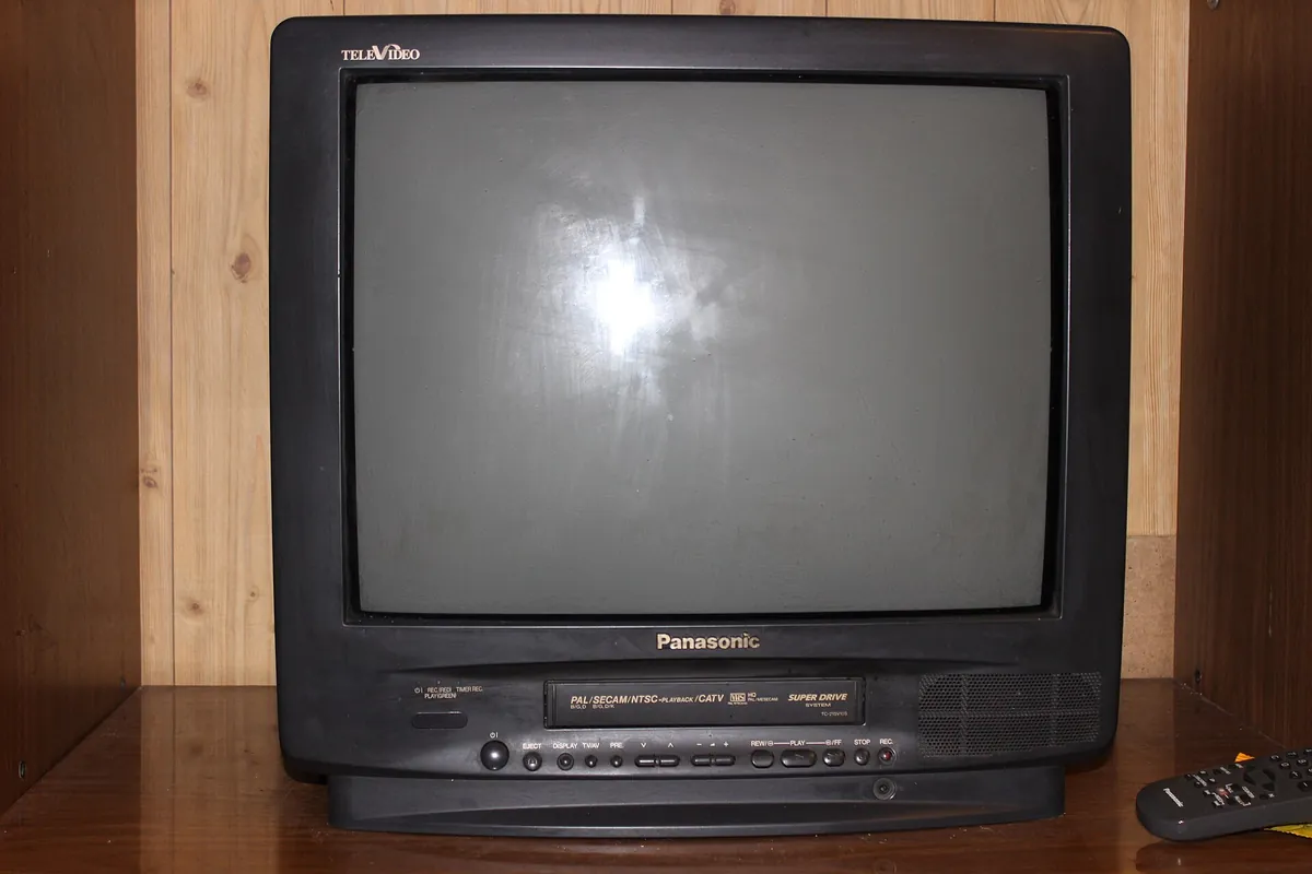 A boxy tube television with a built-in video cassette recorder standing on a wooden shelf, with its remote control beside it.