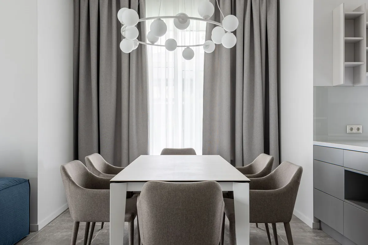 A rectangular six-seat dining table with upholstered chairs under a globe pendant light in a modern apartment, beside a grey kitchen run.