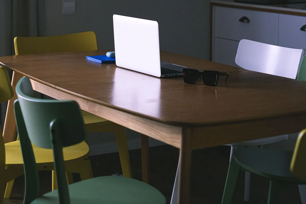 A wooden dining table surrounded by mismatched yellow, green and white chairs, with a laptop open on the table top.
