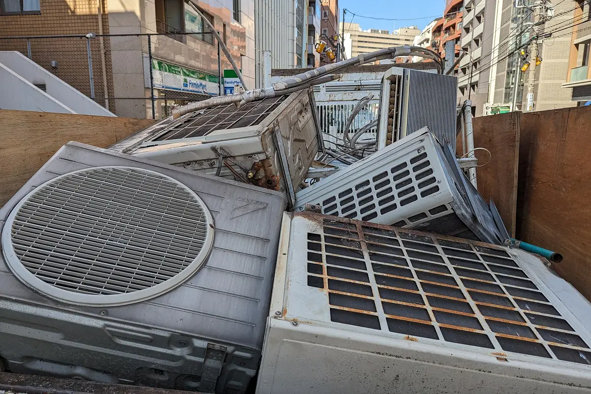 Several old outdoor air conditioner units and window units stacked inside a waste container during a building renovation.