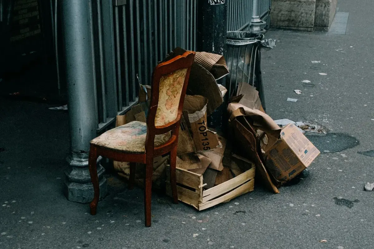 An upholstered dining chair with a worn floral seat left on a pavement beside flattened cardboard, a wooden crate and a street bin.