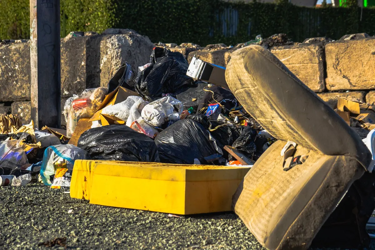 A discarded foam mattress propped against a wall beside a pile of black bin bags, cardboard and a yellow crate at the roadside.