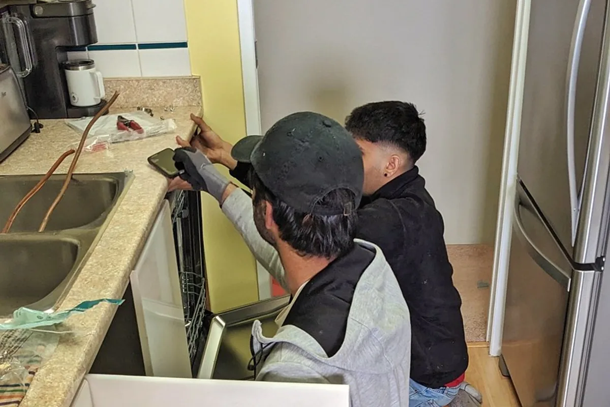 Two workers crouched beside a kitchen sink unit easing a dishwasher out from under the worktop, with its white cabinet door panel removed and propped on the floor.