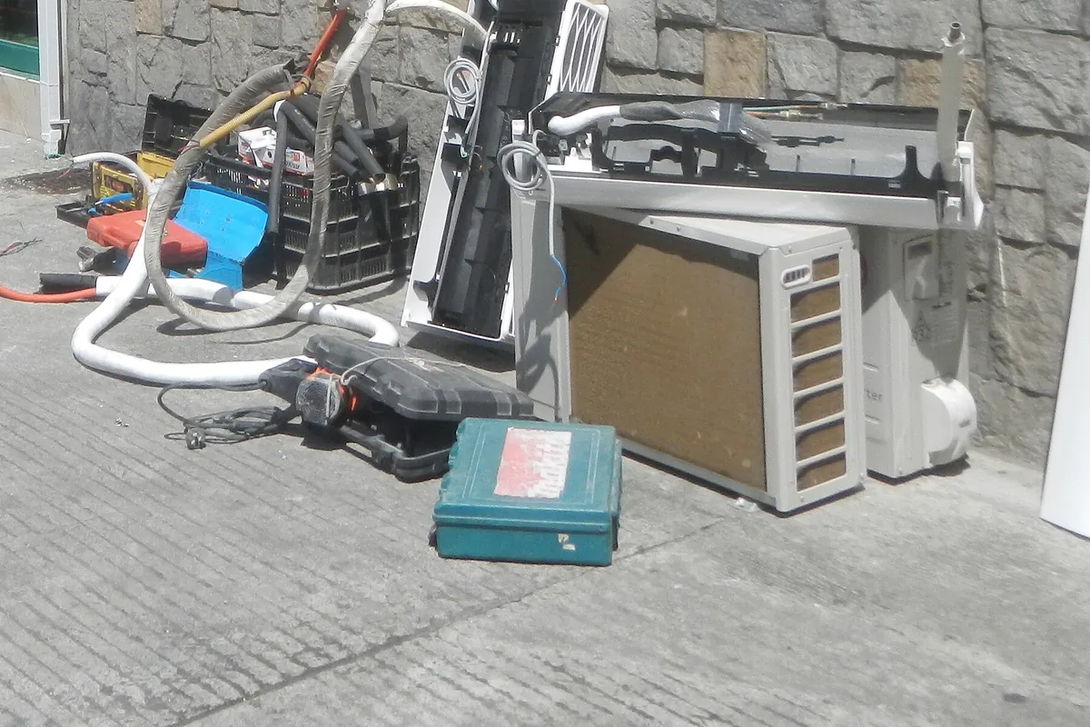 A removed outdoor condenser, an indoor unit casing, coiled insulated pipework and tool cases resting on a pavement after an air conditioner was taken down.