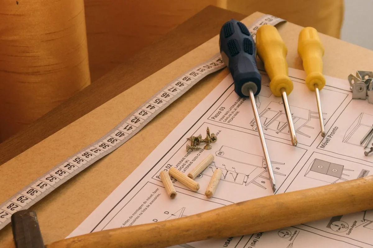 Wooden dowels, coarse screws, three screwdrivers, a tape measure and a hammer resting on an assembly instruction sheet on top of a part-built board carcass.