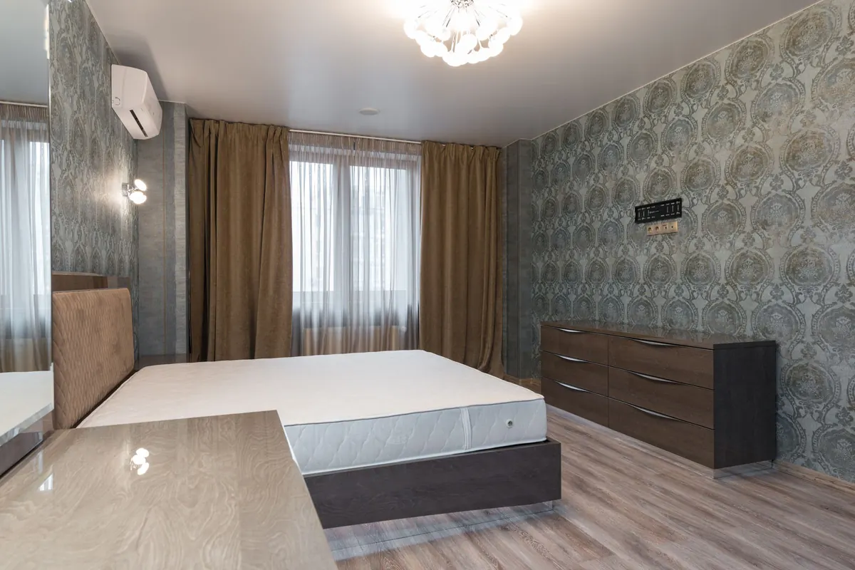 A wide six-drawer chest standing against a patterned wall in an apartment bedroom, beside a bed stripped back to a bare mattress.