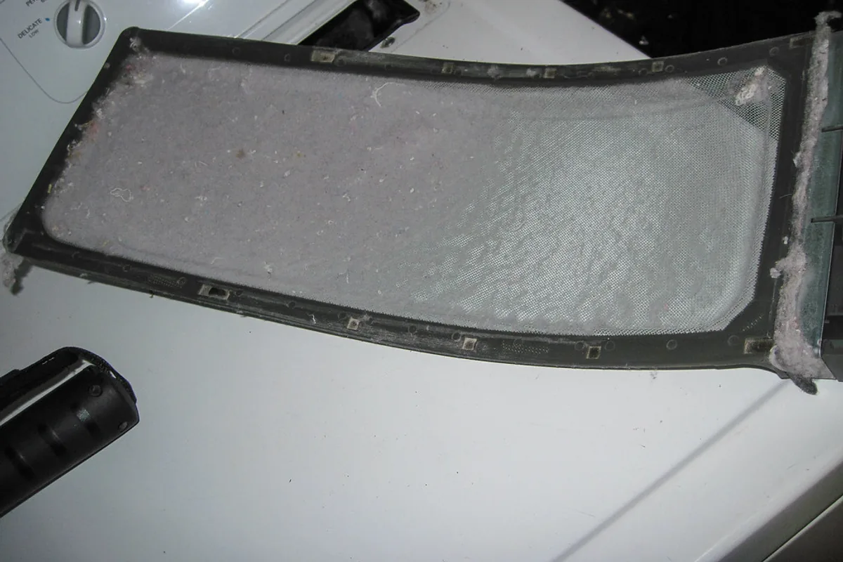 A rectangular tumble dryer lint filter lying on top of a machine, its mesh completely covered by a thick grey mat of accumulated lint.