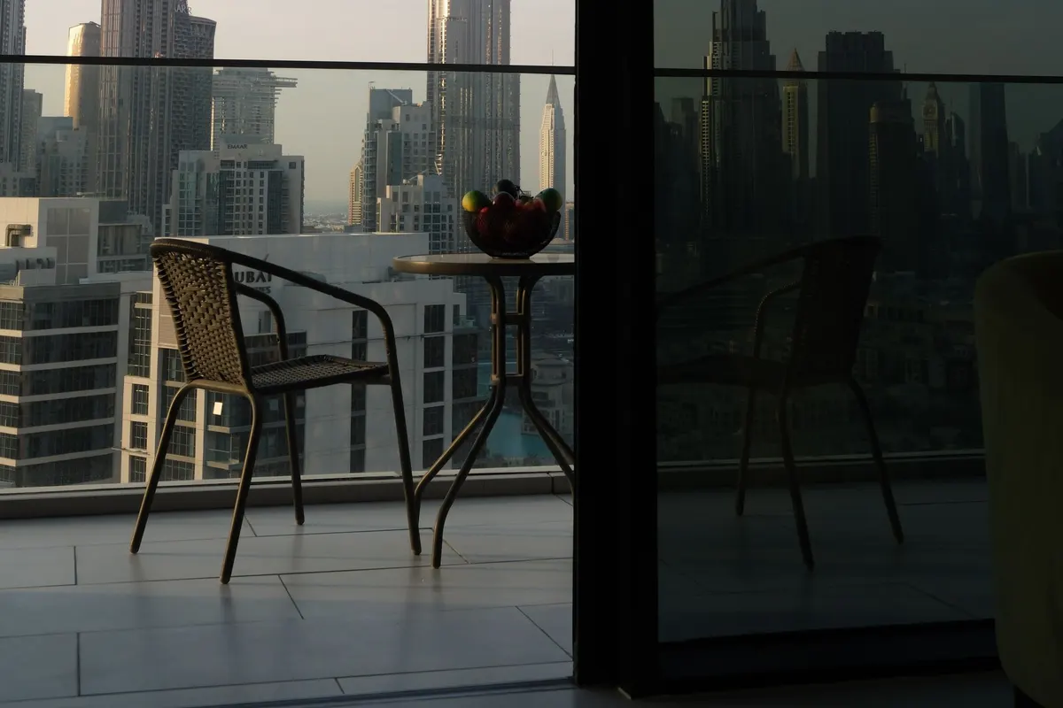 A small table and two chairs on a tiled apartment balcony in Dubai, seen from inside through an open sliding glass door with the city's towers beyond.