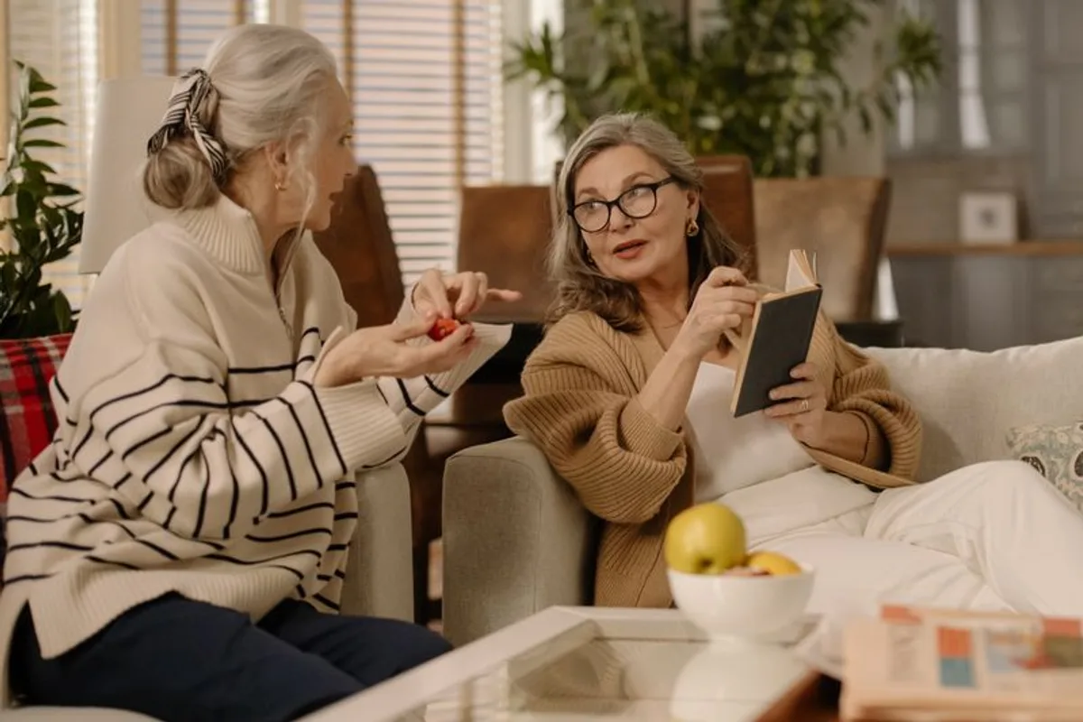 Two older women sitting together on a sofa in a bright living room, talking, with a bowl of fruit on the table in front of them.