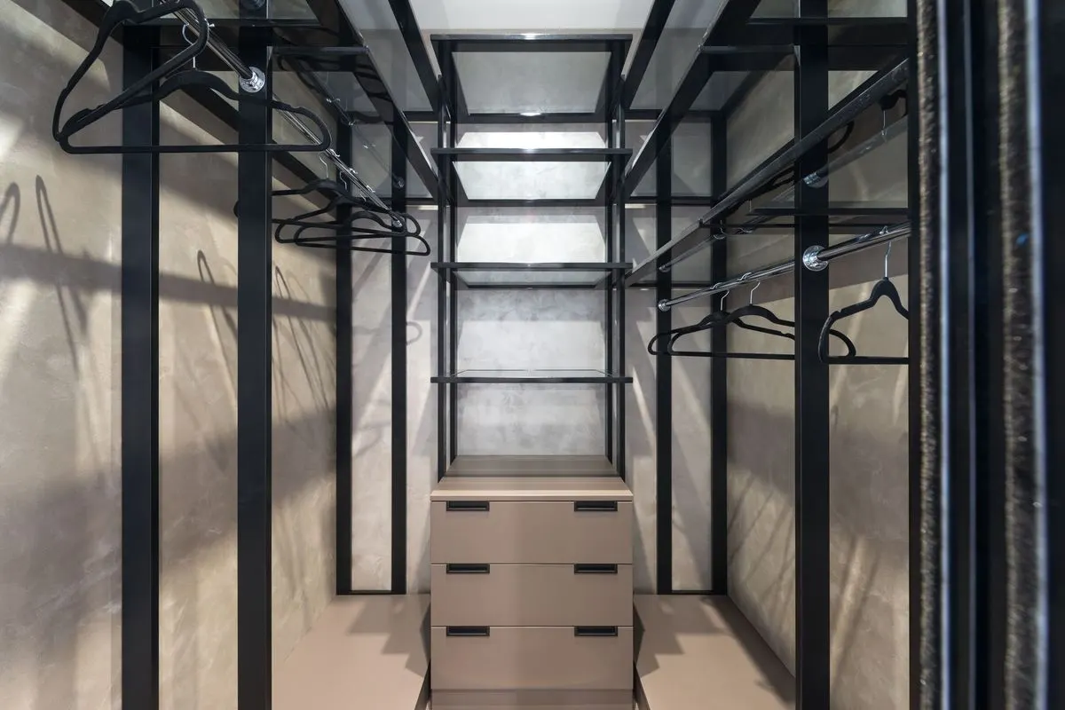 An emptied walk-in wardrobe with black fitted rails and glass shelves along both walls, and a small freestanding chest of drawers standing in the middle.