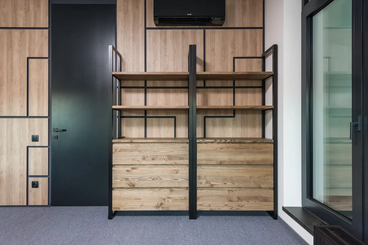 An empty freestanding shelving unit with a black steel frame, timber shelves and drawers below, standing against a panelled wall under a wall-mounted air conditioner.