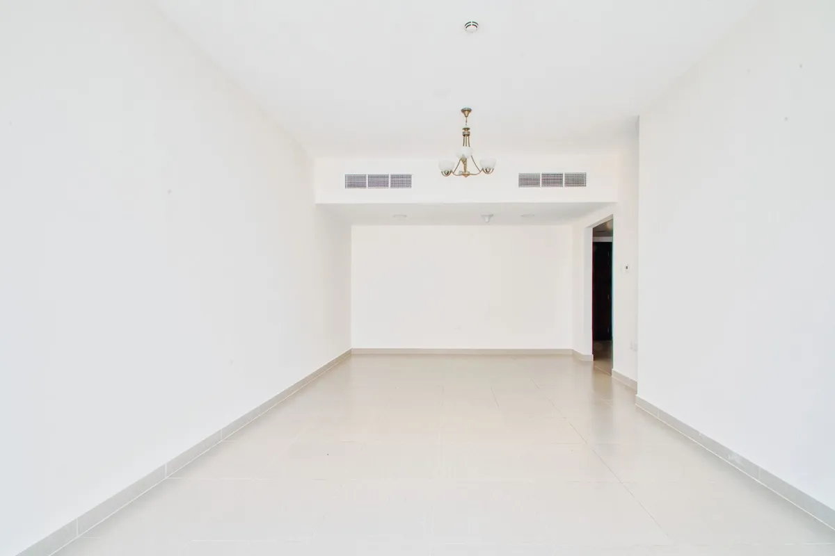 An empty white-walled apartment living room with pale floor tiles, ceiling air-conditioning grilles and a small chandelier, cleared between tenants.