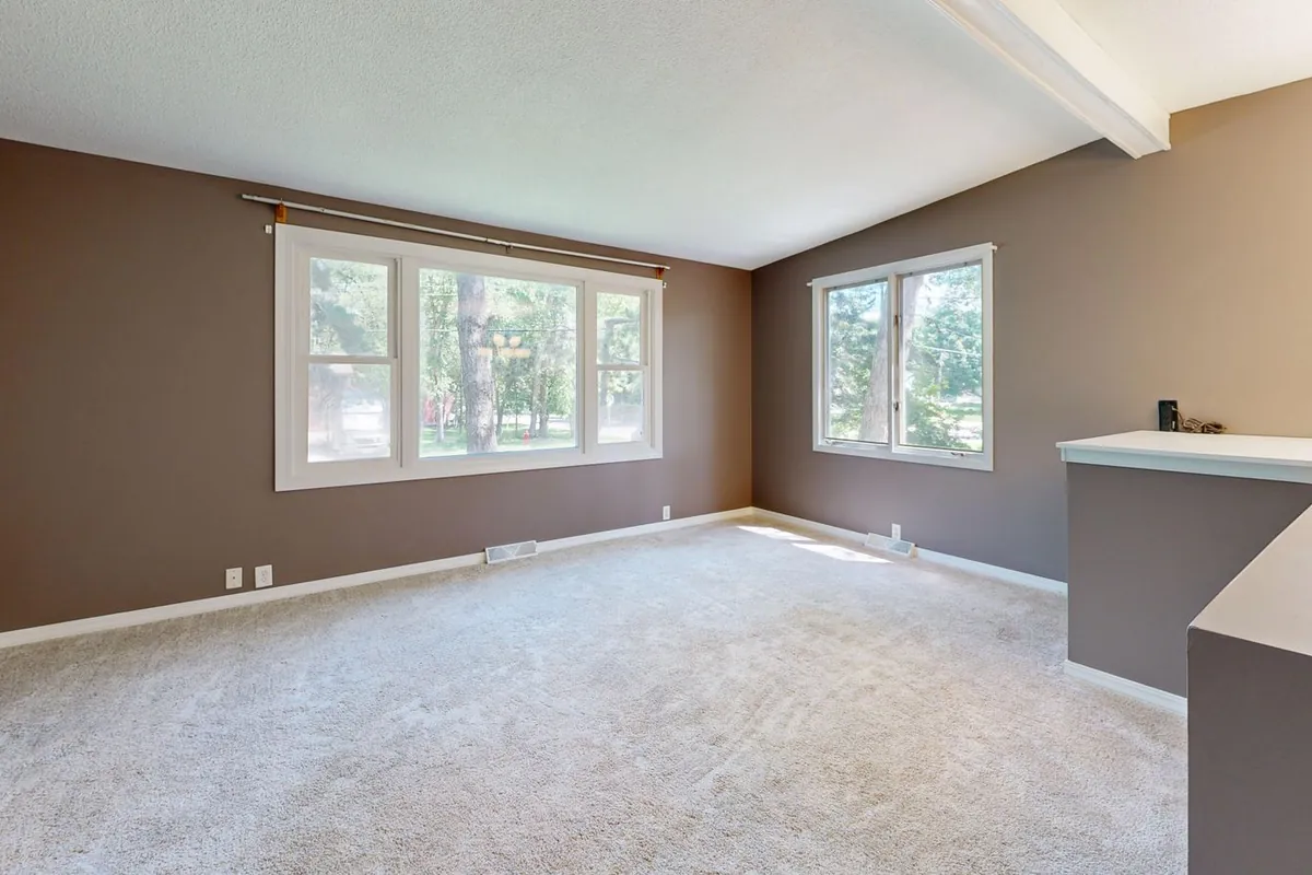 An empty living room with beige wall-to-wall carpet running under the skirting boards to the corners, lit by two windows.