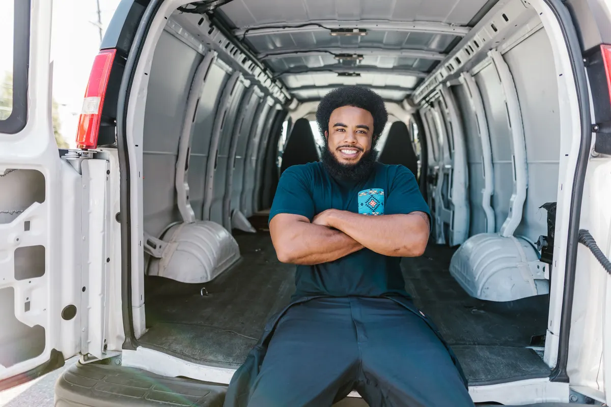 A worker sitting in the completely empty cargo bay of a white van with both rear doors open.