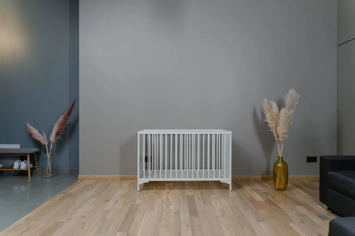 An empty white wooden cot with no mattress standing alone against a grey wall on a pale wooden floor in a modern apartment.