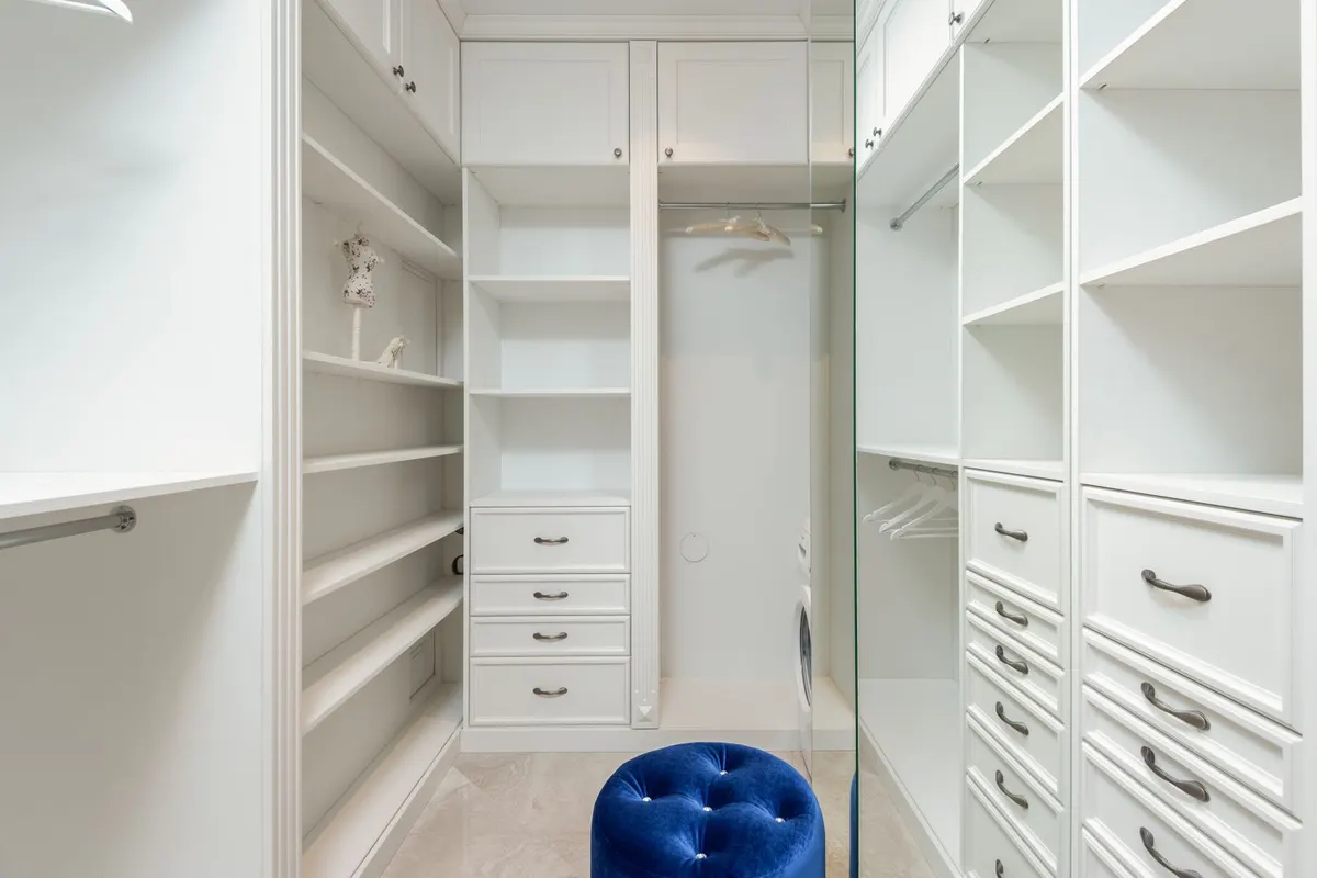 The interior of a white walk-in wardrobe with empty shelves, hanging rails and banks of drawers, with a blue velvet stool on the floor.
