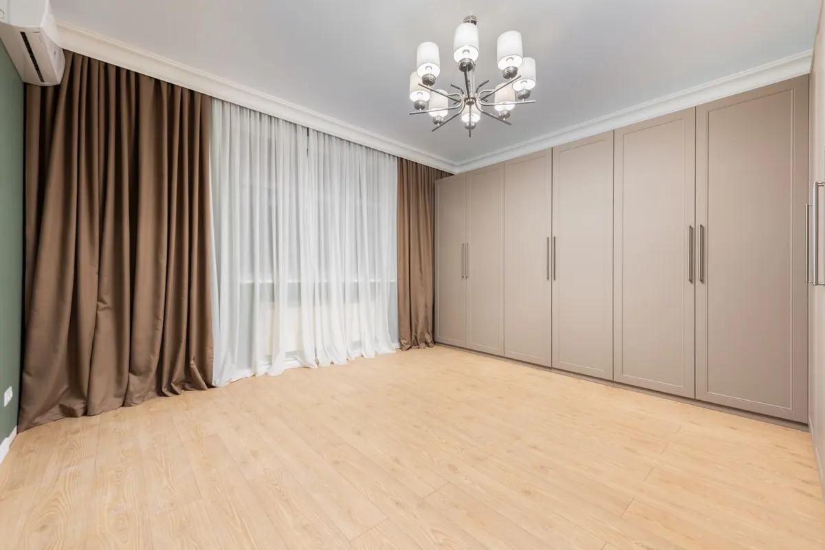 A run of floor-to-ceiling fitted wardrobe doors along the wall of an empty bedroom with wooden flooring and a wall-mounted air conditioning unit.