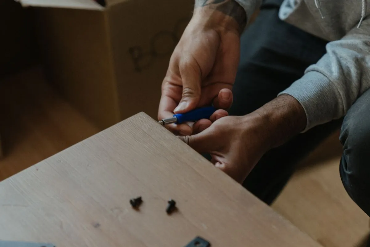 A person driving a fitting into the edge of a laminated furniture panel with a hex key, with corner brackets and loose screws lying on the board and a moving carton behind.