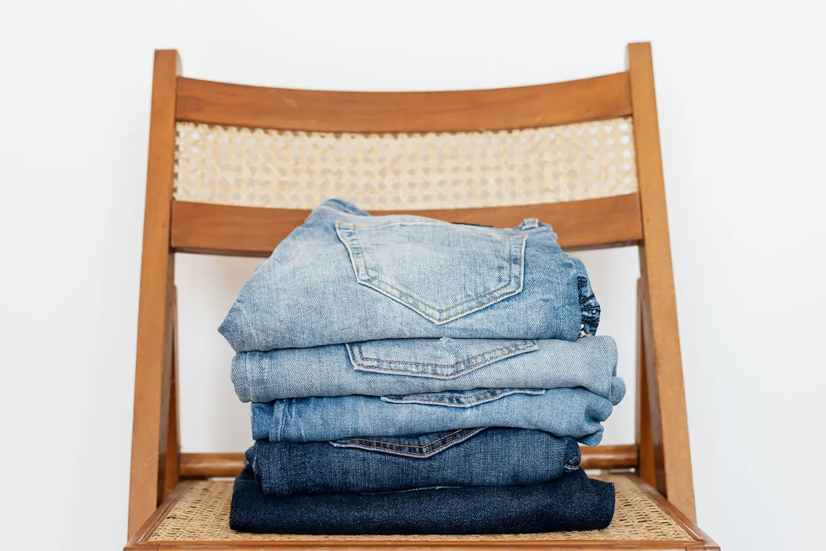 A wooden folding chair with a caned seat and back holding a neat stack of five pairs of folded jeans, against a white wall.