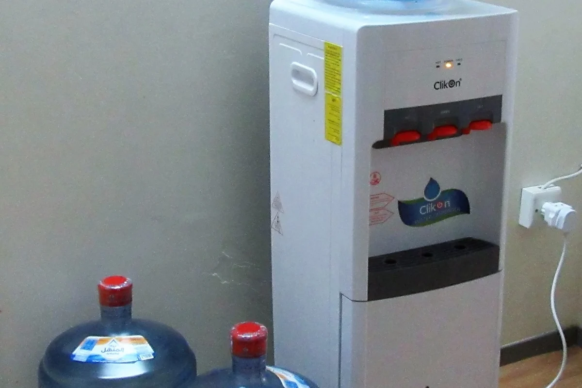 A freestanding white water dispenser unit standing next to five-gallon bottled water jugs ready for removal
