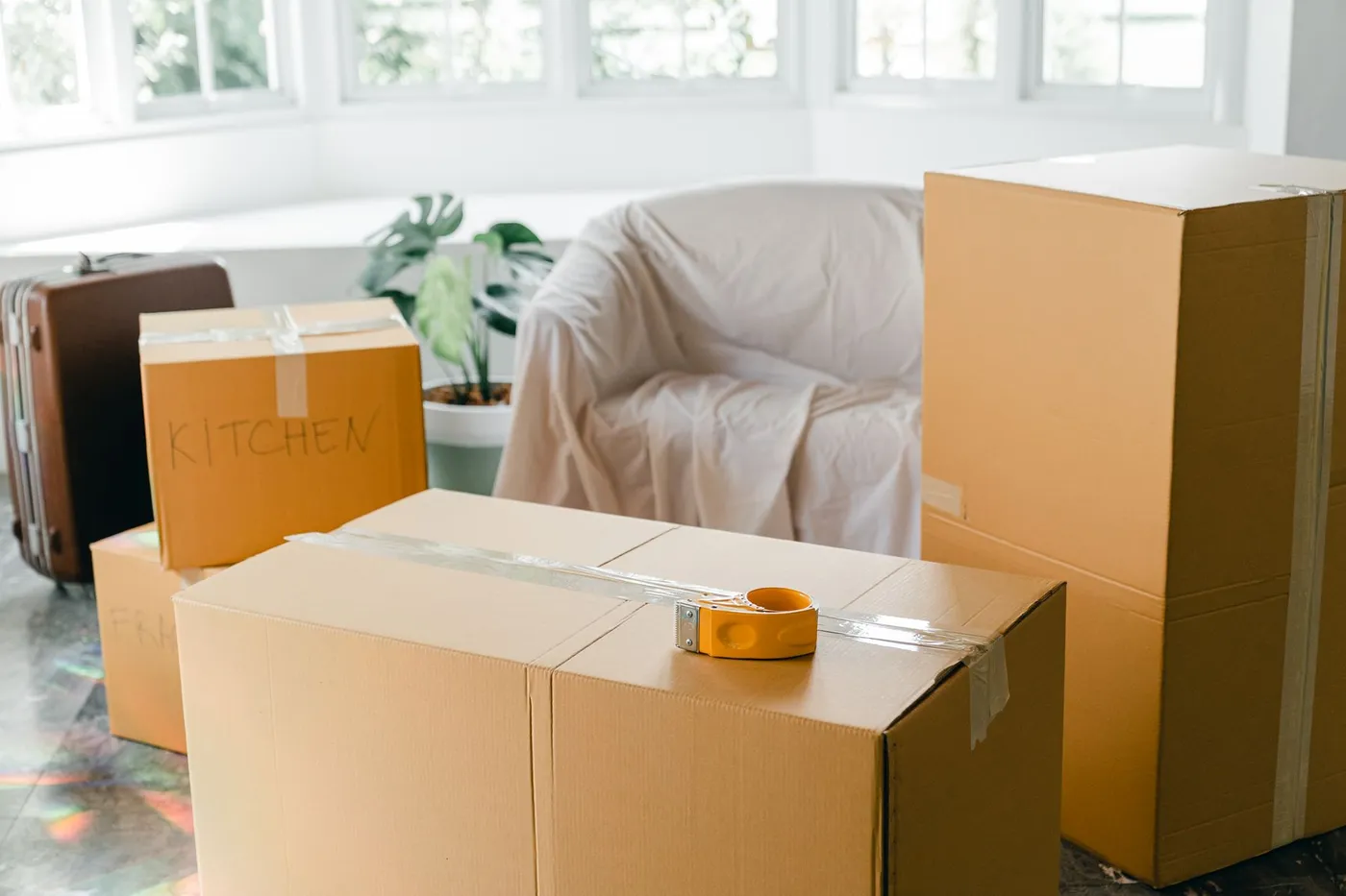 Cardboard boxes labelled by room beside a dust-sheeted armchair and a suitcase in a bright, half-emptied living room.