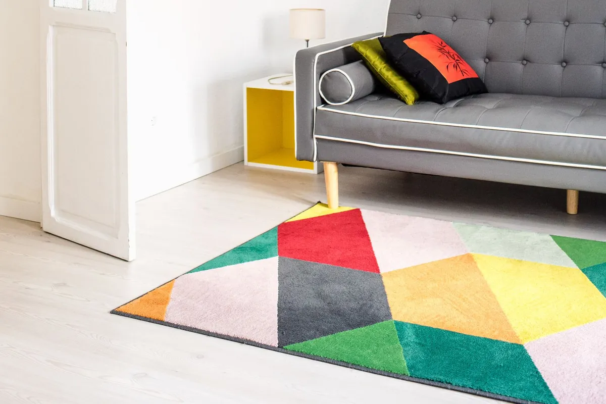 A large geometric area rug in red, green, yellow and grey covering the pale wooden floor of a bright living room in front of a grey sofa.