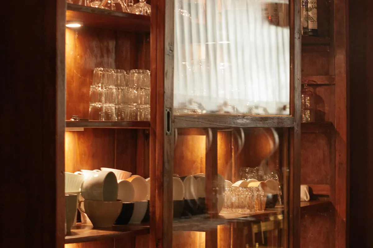 A tall wooden display cabinet with sliding glass doors, lit from inside and filled with stacked tumblers, bowls and bottles.