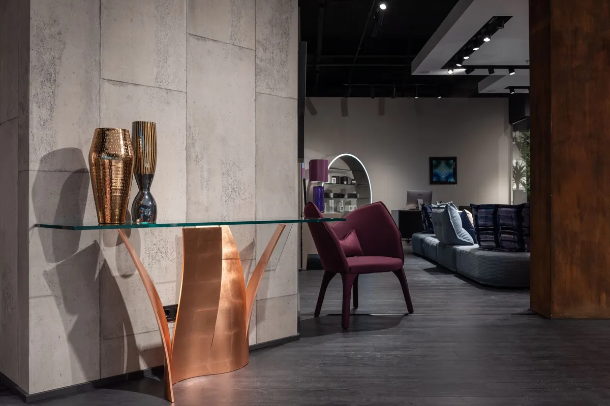A long clear glass tabletop resting on a sculptural copper base against a concrete wall, with the polished edge of the glass sheet clearly visible.