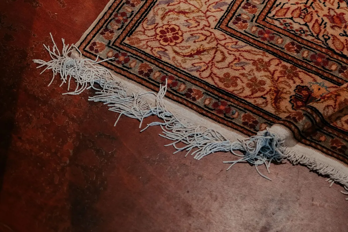 The corner of a finely patterned wool rug on a polished floor, with pale fringe threads running out of the end of the woven border.