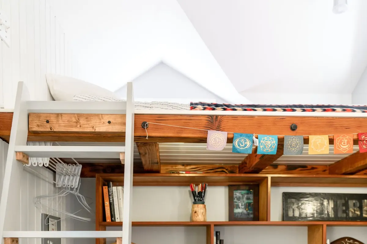 The upper platform of a high sleeper bed resting on a heavy timber beam, with a white ladder at one end and a run of built-in wooden shelving fitted into the space beneath it.