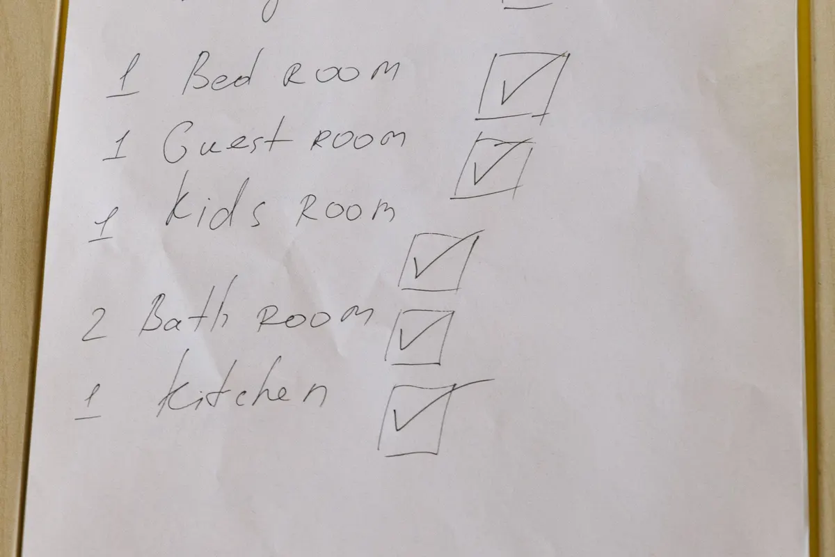A handwritten room-by-room checklist on a clipboard listing bedroom, guest room, kids room, bathrooms and kitchen with ticked boxes.