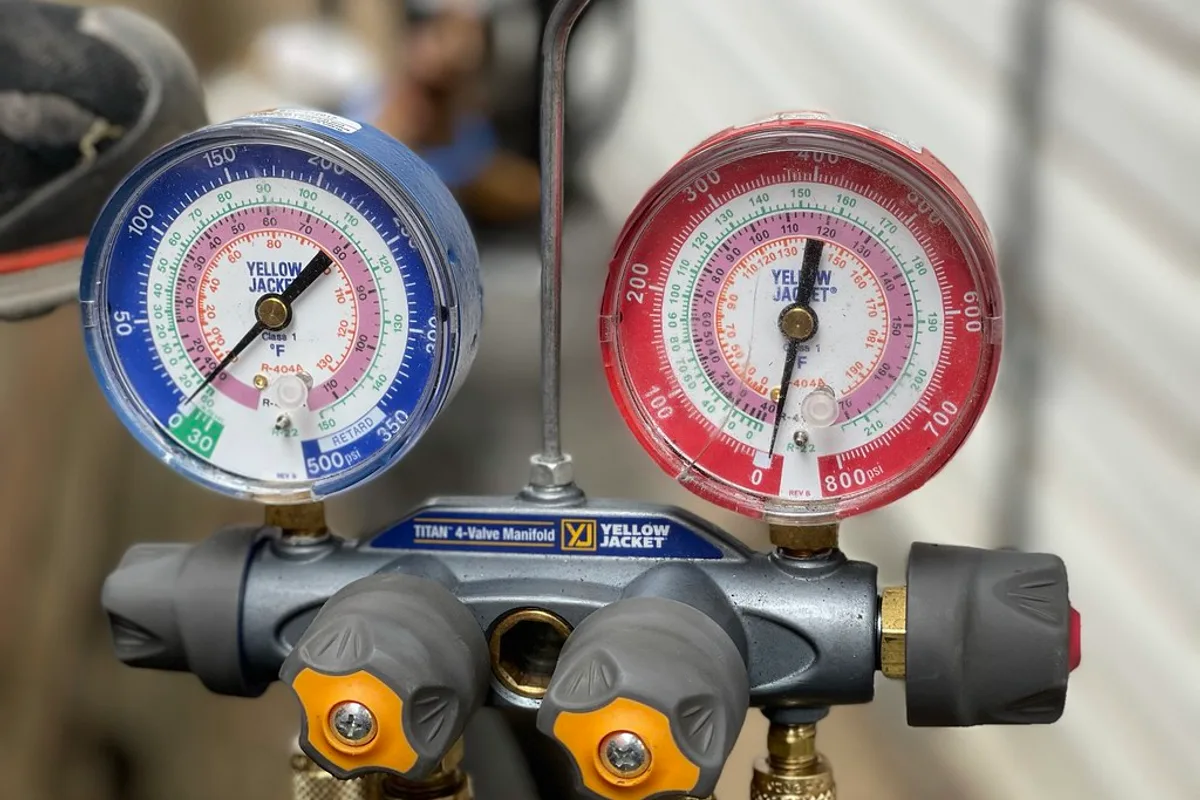 A technician's manifold gauge set with dual pressure dials connected to an air conditioning refrigerant line.