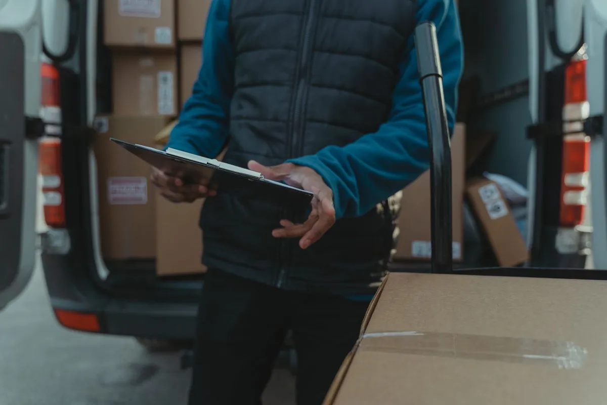 A worker in a blue fleece and dark body warmer holding a clipboard beside the open rear doors of a van loaded with cardboard boxes.