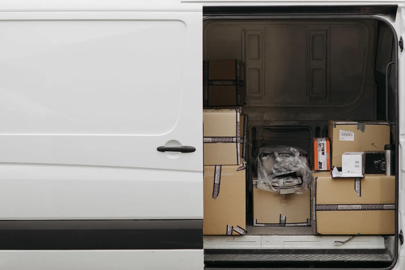 A white van with its side door slid open, the cargo bay part-filled with stacked cardboard boxes and a wrapped item.