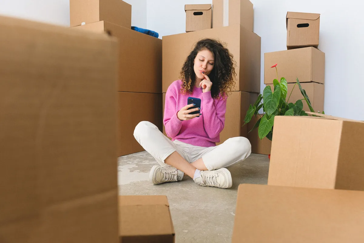 A woman sitting cross-legged among moving boxes, looking thoughtfully at her phone while messaging.