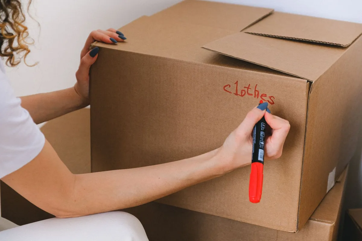 A person writing the word 'clothes' on a cardboard box with a red marker before collection.