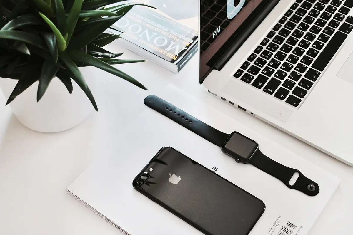 A laptop, a smartphone and a smartwatch laid out on a white desk beside a potted plant and a stack of magazines.
