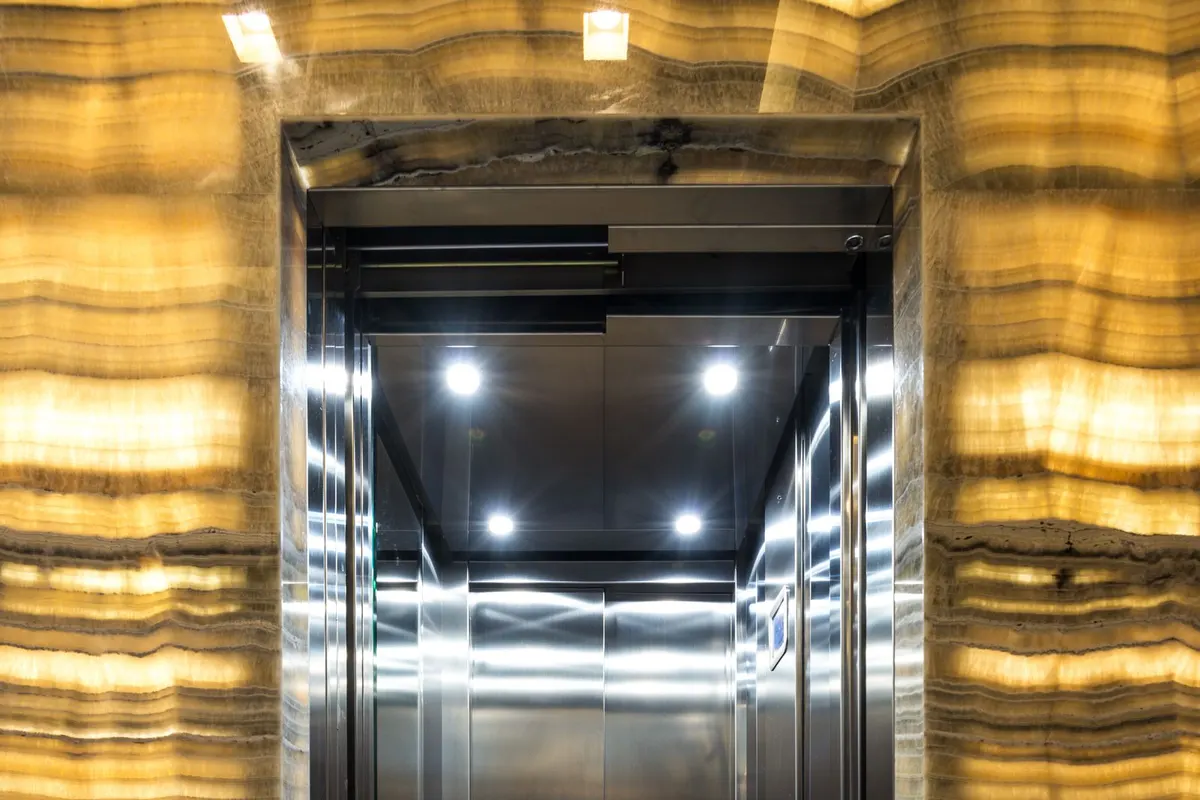 An open stainless steel lift car with mirrored interior panels, set into a backlit stone wall in a residential tower lobby.