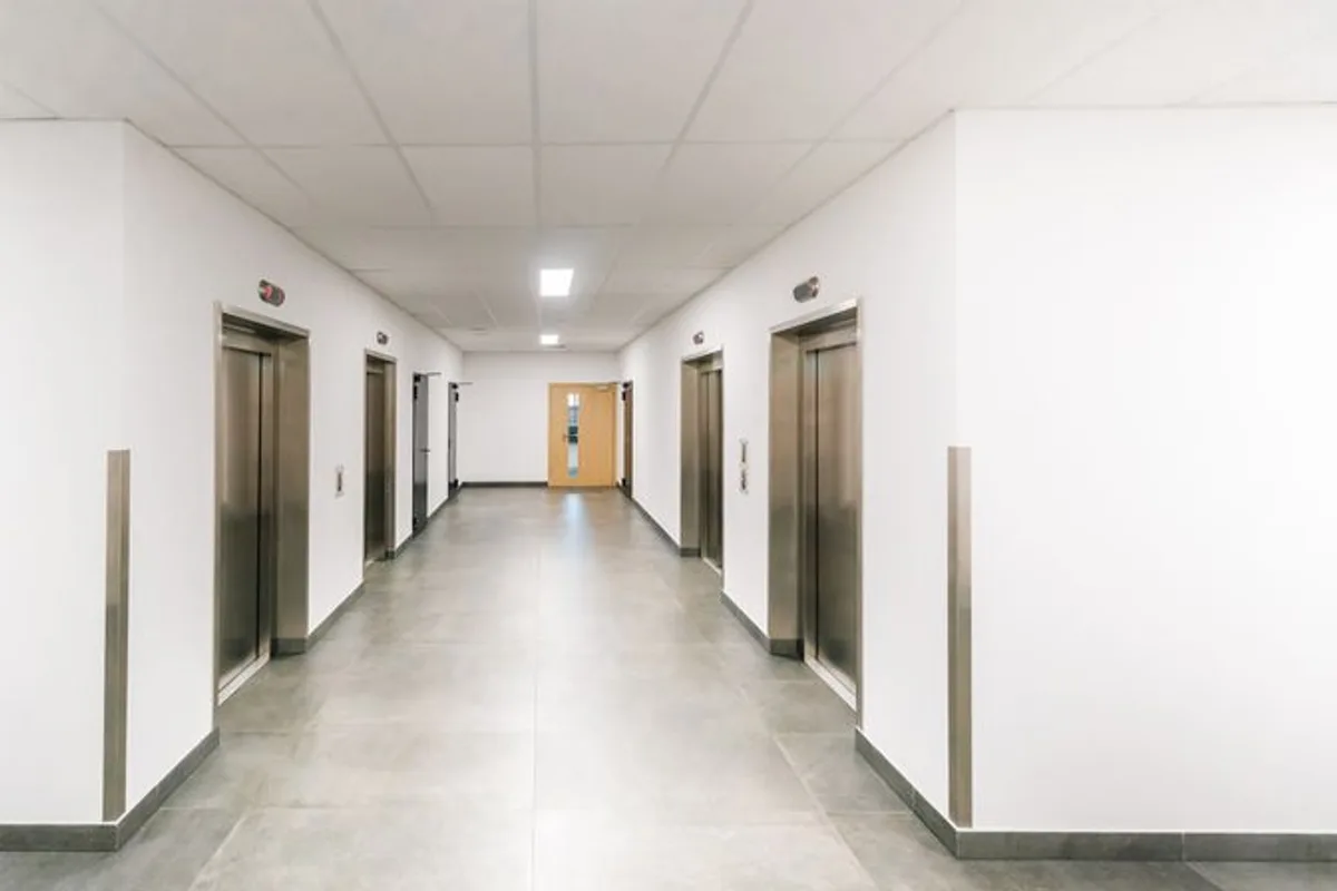 A bright corridor in a modern building with two banks of stainless steel lift doors facing each other.