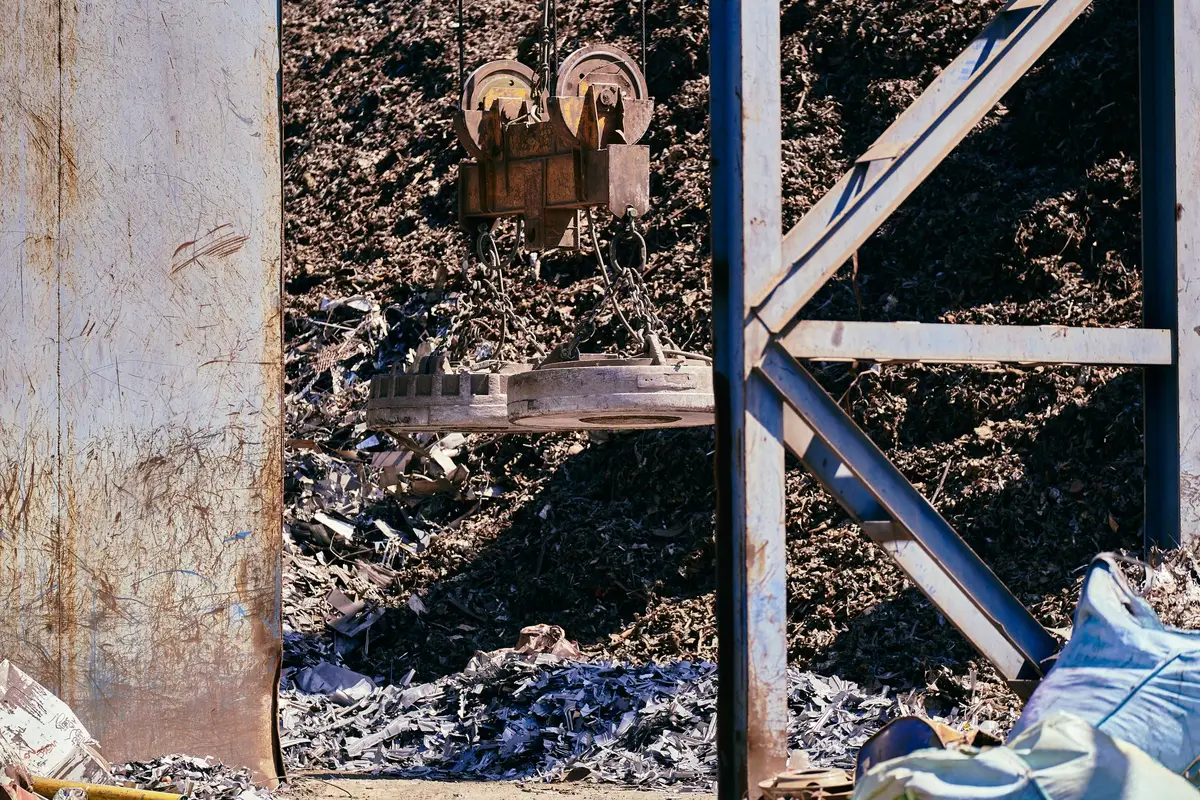 A circular lifting magnet hanging on chains above a large pile of shredded scrap metal in an outdoor recycling yard.
