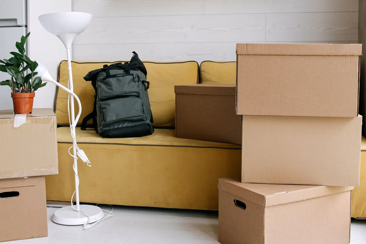 A living room part-way through a move, with a yellow sofa, a backpack and stacks of packed cardboard boxes on a pale floor.